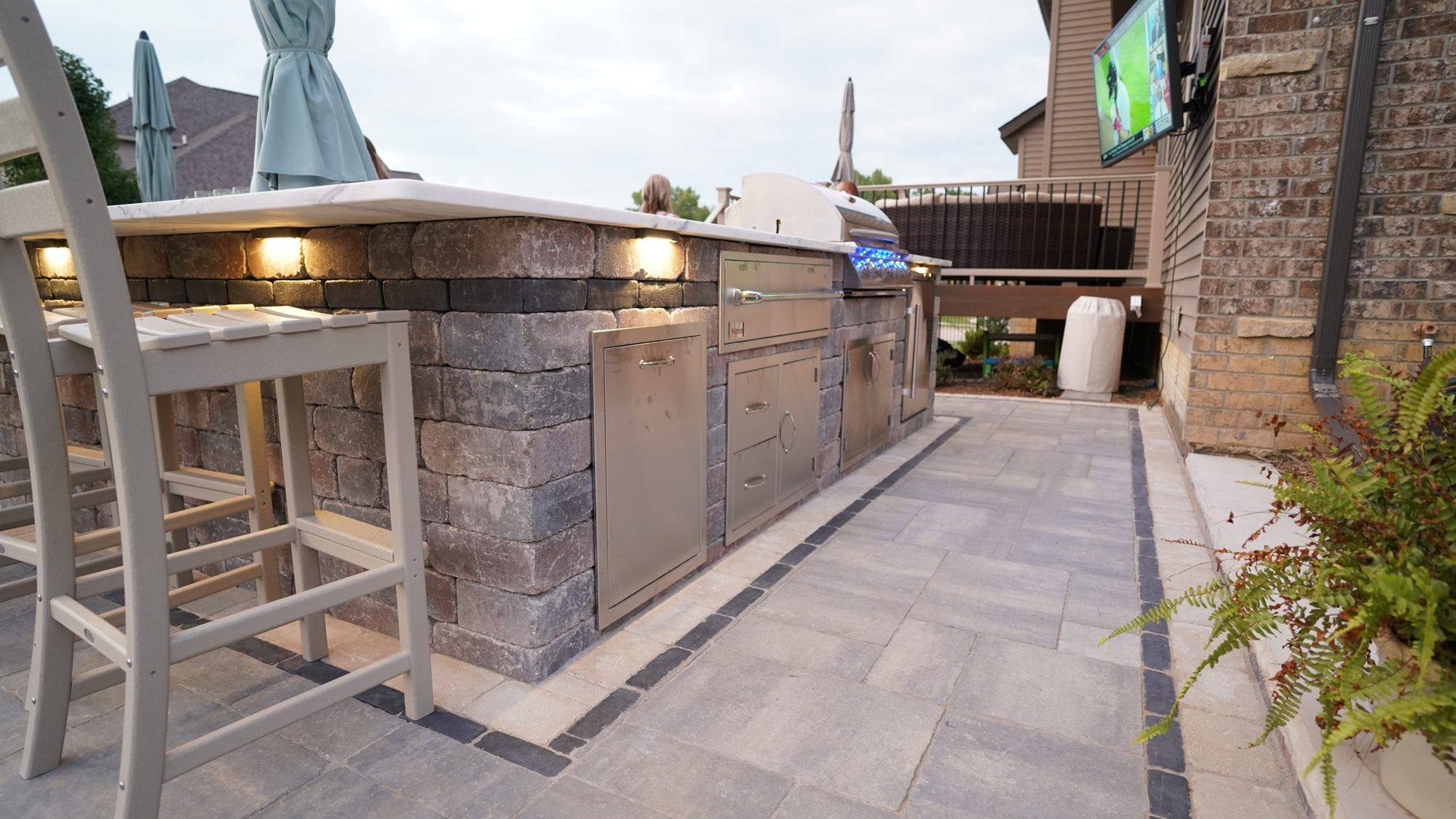 Long outdoor kitchen with bar stools and under-counter lighting on a paver patio